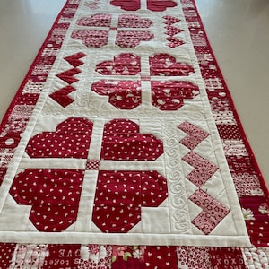 May include: A white table runner with a red and white patchwork design featuring hearts and flowers. The runner has a scalloped edge and is quilted with white thread.