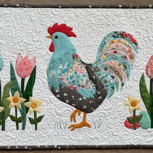 Può includere: Un arazzo trapuntato raffigurante un gallo colorato con cresta rossa e corpo turchese. Il gallo è circondato da tulipani e narcisi. Lo sfondo è bianco con un motivo a spirale e il bordo è grigio scuro con piccoli accenti floreali.