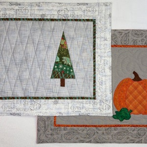 May include: Two quilted placemats with embroidered designs. The first placemat features a green and brown Christmas tree with white accents. The second placemat features a brown and orange pumpkin with green leaves. Both placemats have a white background with grey and white patterned borders.