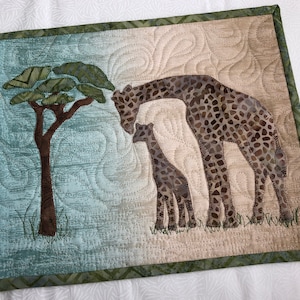 May include: A quilted wall hanging featuring a giraffe and calf under a tree. The giraffes have brown and tan spots, and the tree has green leaves. The background is a mix of blue and tan, with a green border.