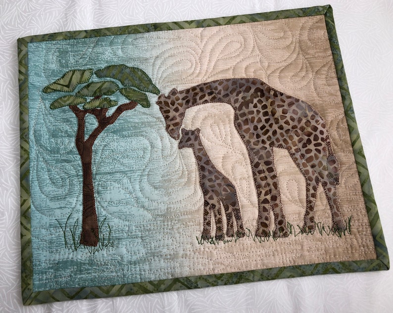 May include: A quilted wall hanging featuring a giraffe and calf under a tree. The giraffes have brown and tan spots, and the tree has green leaves. The background is a mix of blue and tan, with a green border.