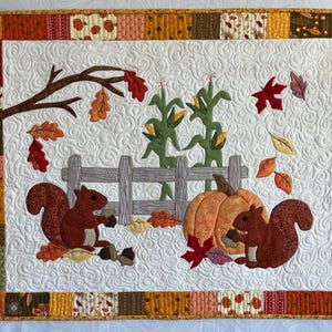 Może przedstawiać: Pikowany obraz na ścianę o tematyce jesiennej. Projekt przedstawia dwa wiewiórki, dynię, kolby kukurydzy i spadające liście. Obramowanie wykonane jest z różnych wzorzystych tkanin w jesiennych kolorach. Tło jest białe z pikowanym wzorem.