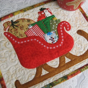May include: A red and brown quilted Christmas coaster featuring a red sleigh filled with presents and a snowman. The coaster has a white background with a floral pattern.