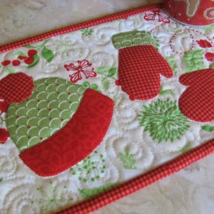 May include: A white quilted pot holder with a red and green winter theme. The design features a red and white gingham mitten, a red and white gingham hat, and green leaves. The pot holder has a red and white gingham border.