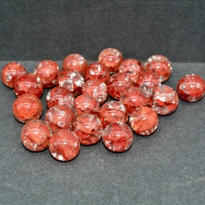May include: A collection of red glass beads with a textured, faceted appearance. The beads are round and scattered on a dark gray surface, creating a visually appealing display. These beads could be used for jewelry making or other craft projects.
