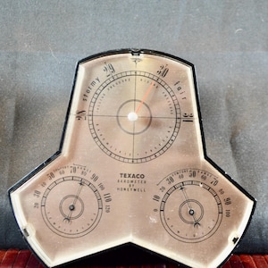 May include: A black-framed Texaco Barometer by Honeywell with a three-part display. The top section shows barometric pressure with a needle, while the two lower sections display temperature and humidity levels with individual needles on a beige face.
