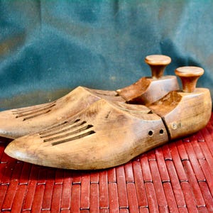 May include: A pair of vintage wooden shoe forms, showcasing a warm, natural wood tone. Each form has a rounded toe, ventilation slots, and a knob at the heel. They rest on a red bamboo mat against a dark teal backdrop.