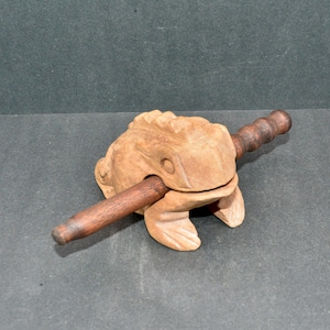 May include: A wooden frog-shaped musical instrument, with a dark brown wooden stick. The frog is light brown with carved details. The stick is inserted through the frog's mouth. A percussion instrument.