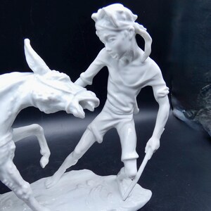 Może przedstawiać: Biała porcelanowa figurka przedstawiająca osobę trzymającą kij i osła. Osoba ma opaskę na głowie i koszulę z kr&oacute;tkim rękawem. Osioł stoi na tylnych nogach i ma głowę odwr&oacute;coną w bok.