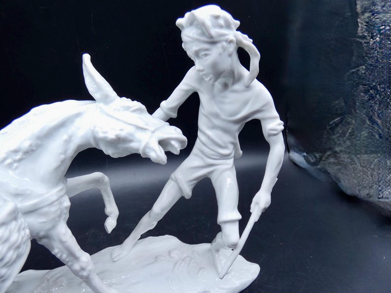 Może przedstawiać: Biała porcelanowa figurka przedstawiająca osobę trzymającą kij i osła. Osoba ma opaskę na głowie i koszulę z kr&oacute;tkim rękawem. Osioł stoi na tylnych nogach i ma głowę odwr&oacute;coną w bok.