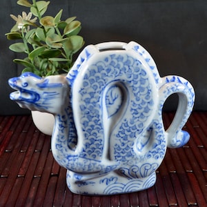 May include: A white and blue ceramic teapot in the shape of a dragon. The teapot features a detailed dragon design with blue accents. The handle and spout are integrated into the dragon's form. Decorative waves are at the base.