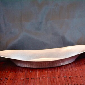 May include: A long, shallow, silver-colored decorative bowl. The bowl has a curved, boat-like shape with a wide opening and gently sloping sides. It sits on a dark red bamboo mat against a dark background.