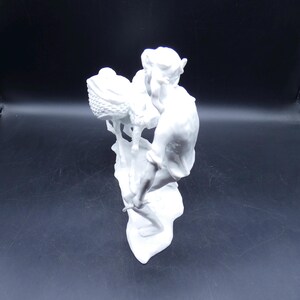 Może przedstawiać: Biała porcelanowa figurka przedstawiająca osobę trzymającą koszyk z dzieckiem w środku. Figurka stoi na podstawie z fakturowaną powierzchnią.