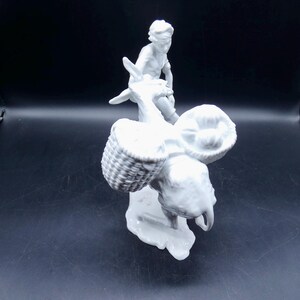 Może przedstawiać: Biała porcelanowa figurka osoby jadącej na osiołku z dwoma koszykami na grzbiecie.