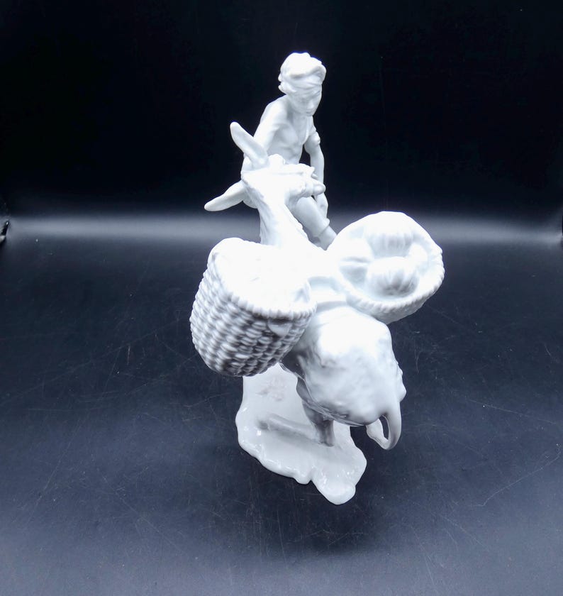 Może przedstawiać: Biała porcelanowa figurka osoby jadącej na osiołku z dwoma koszykami na grzbiecie.