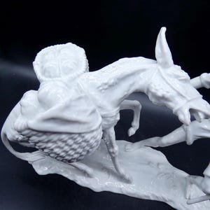 Może przedstawiać: Biała porcelanowa figurka osła niosącego kosz z produktami, z mężczyzną ciągnącym za wodze osła.