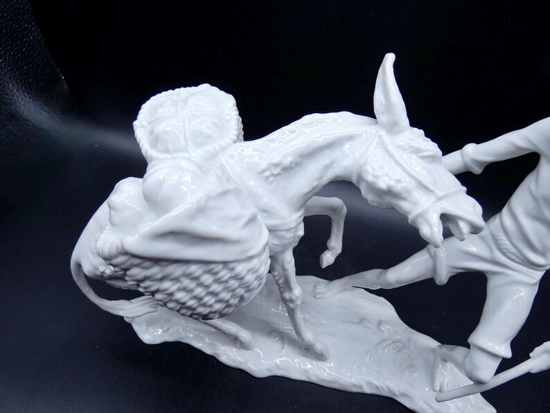Może przedstawiać: Biała porcelanowa figurka osła niosącego kosz z produktami, z mężczyzną ciągnącym za wodze osła.