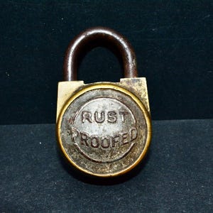 May include: An antique padlock with a dark brown shackle and a brass-colored body. The face of the lock is circular, with the words "RUST PROOFED" embossed in a circular pattern. The lock is set against a dark background.