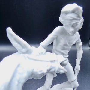 Może przedstawiać: Biała porcelanowa figurka chłopca prowadzącego osła. Chłopiec ma na sobie prostą koszulę i opaskę na głowie. Osioł patrzy w bok.