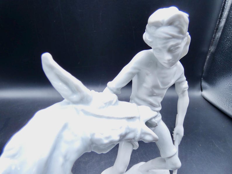 Może przedstawiać: Biała porcelanowa figurka chłopca prowadzącego osła. Chłopiec ma na sobie prostą koszulę i opaskę na głowie. Osioł patrzy w bok.