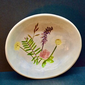 May include: A white ceramic bowl with a hand-painted floral design. The bowl features a cluster of wildflowers, including a pink flower, green fern-like leaves, and yellow and purple blooms. The bowl has a light blue wash.
