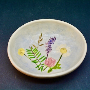 May include: A small, round, off-white ceramic bowl with a hand-painted floral design. The flowers include yellow, purple, pink, and white blooms with green foliage. The bowl sits on a dark blue surface.