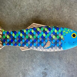 May include: A wooden fish decoration with a blue head and a body covered in blue, green, and gray scales. The tail and fins are light brown with detailed patterns. The fish has a yellow eye.
