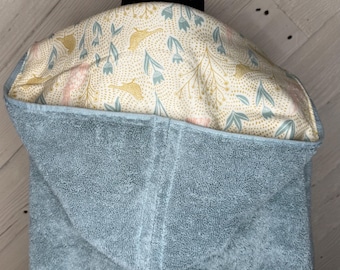 May include: A baby hooded towel in a soft blue hue. The hood's interior showcases a patterned fabric with floral and bird motifs in gold, pink, and blue against a cream backdrop. The towel is crafted from a plush, absorbent material.