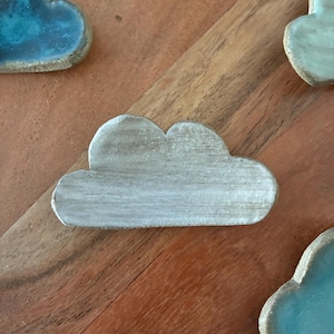 May include: Cloud-shaped ceramic pieces in shades of gray and blue. The cloud-shaped objects are on a wooden surface. The ceramic pieces have a textured appearance and are likely handmade. The pieces are likely decorative.