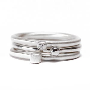 Könnte beinhalten: Ein Stapel aus drei silbernen Ringen mit matter Oberfläche. Der oberste Ring hat einen kleinen, eingelassenen klaren Edelstein, der mittlere Ring eine glatte, polierte Silberkugel und der untere Ring einen kleinen, gebürsteten Silberwürfel.