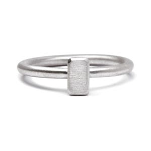 Könnte beinhalten: Ein minimalistischer Silberring mit rechteckigem Stangendesign. Der Ring hat eine schlichte, moderne Ästhetik mit gebürsteter Metalloberfläche. Das Band hat eine glatte, abgerundete Form. Der Ring ist vor weißem Hintergrund.