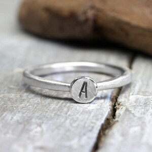 Könnte beinhalten: Ein silberner Ring mit einer runden, strukturierten Scheibe, auf der der Buchstabe "A" eingraviert ist.