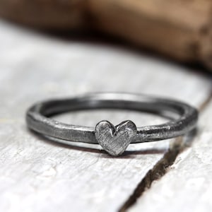 Könnte beinhalten: Ein silberner Ring mit einem herzförmigen Design. Der Ring ist aus Metall gefertigt und hat eine raue, strukturierte Oberfläche.