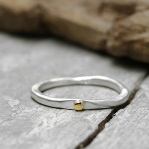 Könnte beinhalten: Ein silberner Ring mit einem strukturierten, organischen Design. Der Ring hat einen kleinen, runden Goldakzent. Der Ring liegt auf einer grauen, strukturierten Oberfläche mit einem Holzelement im Hintergrund.