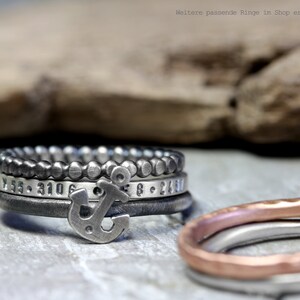 May include: A set of four silver rings with an anchor charm. The rings are stacked on top of each other and have different textures and designs. The top ring is a beaded ring, the second ring has a hammered texture, the third ring has a smooth surface with numbers engraved on it, and the bottom ring is a simple band. The anchor charm is attached to the third ring.