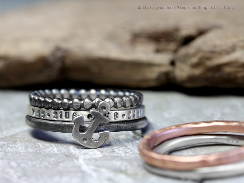 May include: A set of four silver rings with an anchor charm. The rings are stacked on top of each other and have different textures and designs. The top ring is a beaded ring, the second ring has a hammered texture, the third ring has a smooth surface with numbers engraved on it, and the bottom ring is a simple band. The anchor charm is attached to the third ring.