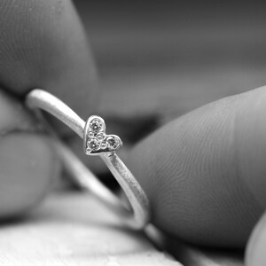 May include: A silver-toned ring featuring a small heart-shaped centerpiece. The heart is adorned with three tiny, clear, circular stones arranged in a triangular pattern. The band of the ring has a matte, textured finish and a simple, rounded design.