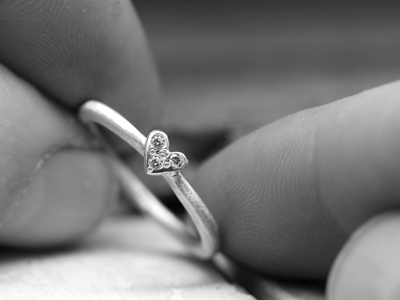 May include: A silver-toned ring featuring a small heart-shaped centerpiece. The heart is adorned with three tiny, clear, circular stones arranged in a triangular pattern. The band of the ring has a matte, textured finish and a simple, rounded design.