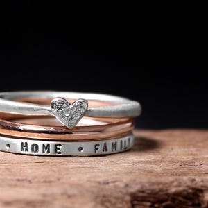 May include: A stack of three rings featuring a silver band with a heart-shaped charm containing small clear stones, a smooth rose gold band, and a silver band stamped with the words FAITH, HOME, and FAMILY separated by small dots.
