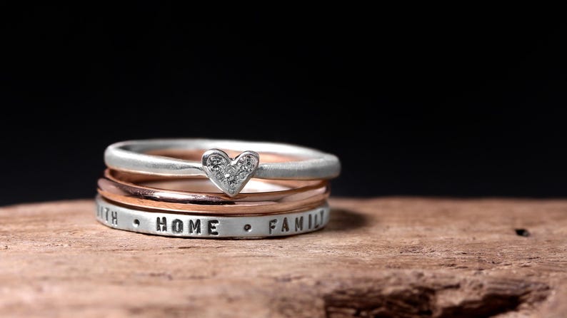May include: A stack of three rings featuring a silver band with a heart-shaped charm containing small clear stones, a smooth rose gold band, and a silver band stamped with the words FAITH, HOME, and FAMILY separated by small dots.