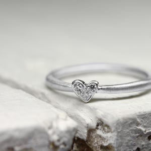May include: A silver-toned ring featuring a small heart-shaped centerpiece. The heart is embellished with five tiny, clear, round-cut stones arranged in a cluster. The band has a smooth, matte finish, creating a subtle contrast with the sparkling stones.