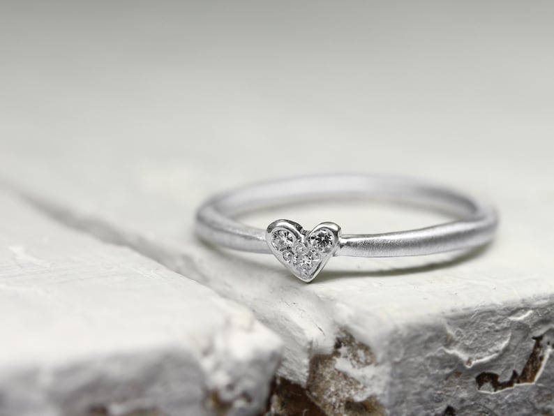 May include: A silver-toned ring featuring a small heart-shaped centerpiece. The heart is embellished with five tiny, clear, round-cut stones arranged in a cluster. The band has a smooth, matte finish, creating a subtle contrast with the sparkling stones.