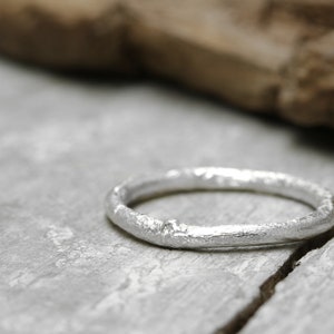 Könnte beinhalten: Ein silberner Ring mit strukturierter Oberfläche. Der Ring ist vor einem grauen Hintergrund mit einem Holzelement im Hintergrund platziert. Der Ring scheint handgefertigt zu sein und hat eine rustikale, organische Ausstrahlung.