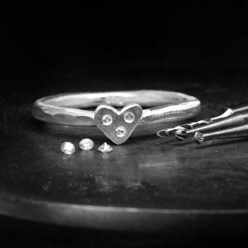 May include: A silver ring featuring a heart-shaped centerpiece with three small, clear, round-cut stones embedded in the surface. Three additional loose, clear, round-cut stones are positioned near the ring, alongside metal jewelry tools.