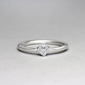 May include: A silver-toned ring featuring a thin, smooth band and a small heart-shaped centerpiece. The heart is set with three tiny, clear, sparkling stones arranged in a triangular pattern, creating a delicate and refined appearance.