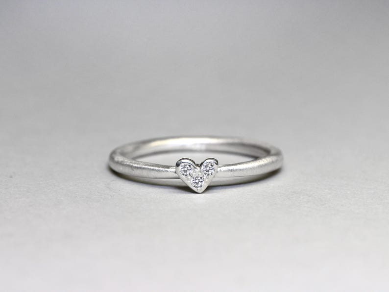 May include: A silver-toned ring featuring a thin, smooth band and a small heart-shaped centerpiece. The heart is set with three tiny, clear, sparkling stones arranged in a triangular pattern, creating a delicate and refined appearance.