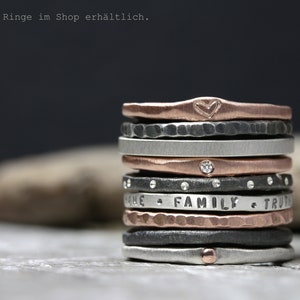May include: A stack of silver, black, and rose gold rings with various textures and engravings. The top ring has a heart engraved on it. The rings are stacked on a gray surface.