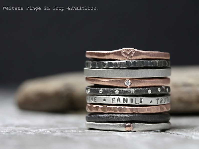 May include: A stack of silver, black, and rose gold rings with various textures and engravings. The top ring has a heart engraved on it. The rings are stacked on a gray surface.