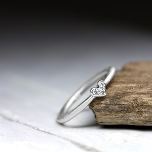 May include: A silver-toned ring featuring a small heart-shaped setting at the top. The heart is embellished with three tiny, clear, sparkling stones arranged in a triangular pattern. The band of the ring is smooth, thin, and has a matte metallic finish.