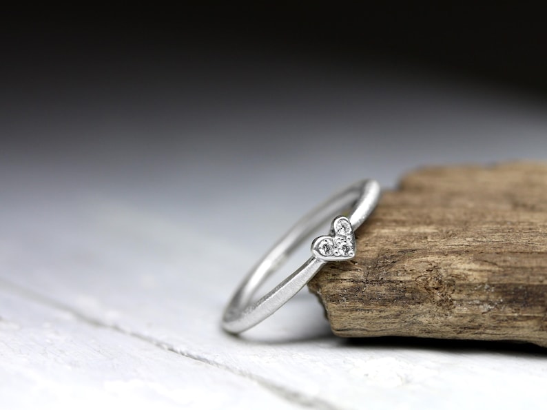 May include: A silver-toned ring featuring a small heart-shaped setting at the top. The heart is embellished with three tiny, clear, sparkling stones arranged in a triangular pattern. The band of the ring is smooth, thin, and has a matte metallic finish.